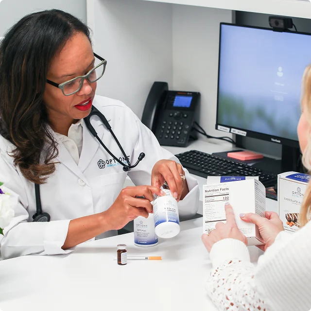 Doctor in a white coat sits at a desk, explaining medication to a patient. There's a computer, phone, and various medicine boxes on the desk.