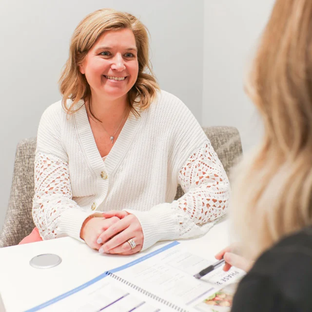 A woman in a white sweater smiles during an office meeting, sitting across a table from another person with a notebook.
