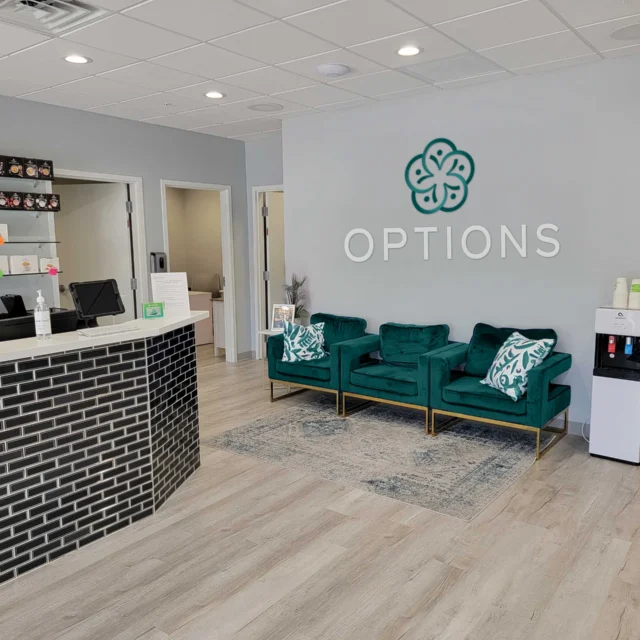 Modern reception area with a black-tiled front desk, green sofas, a water cooler, and "OPTIONS" logo on the wall. Shelves with products are visible behind the desk.