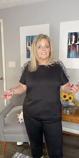 A woman with blonde hair wearing a black shirt and pants stands in a living room near a gray chair, with a small dog sitting on the chair and two paintings on the wall behind her.