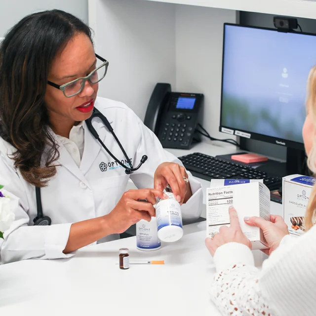 Doctor in a white coat sits at a desk, explaining medication to a patient. There's a computer, phone, and various medicine boxes on the desk.