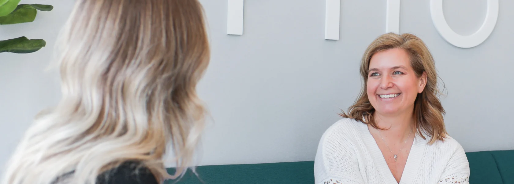 Two women sit on a green couch in conversation; one is facing the camera and smiling, while the other is seen from behind.
