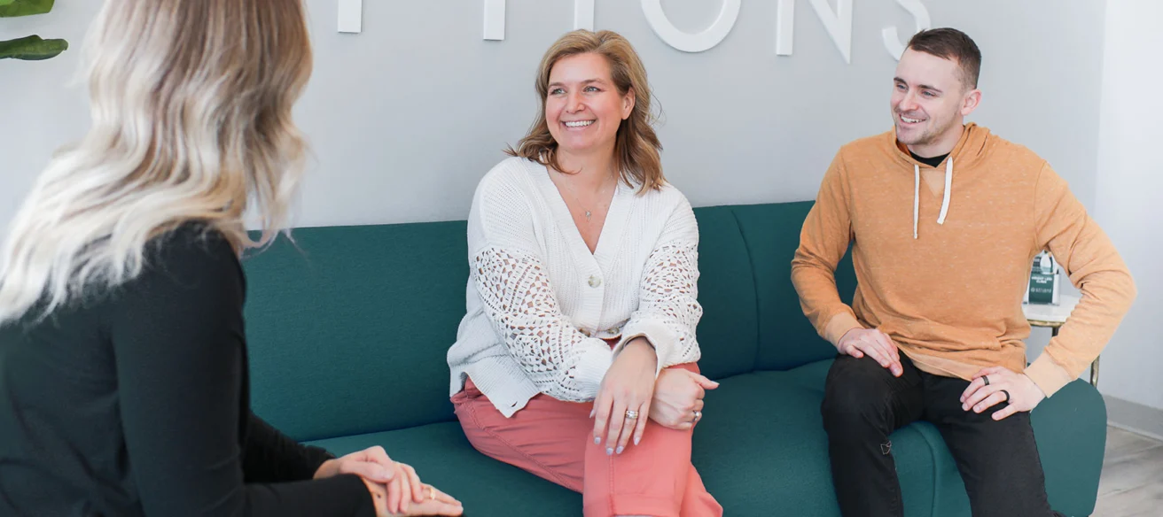 Three people are sitting on a teal couch having a conversation. The woman in the center is smiling, wearing a white sweater and pink pants. The man on the right is in a mustard hoodie.