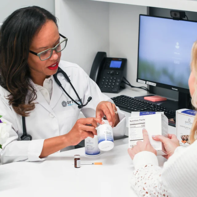 A doctor in a white coat explains medication to a patient sitting across the desk in a medical office, with boxes and a small bottle on the table.