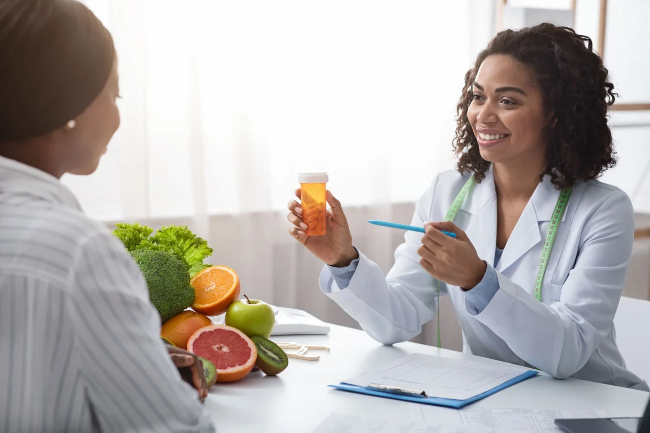 A doctor holding a pill bottle and pen sits across from a patient. Fresh fruits and vegetables are on the table.