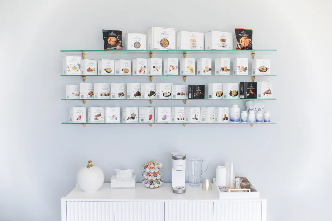 A minimalist coffee station with a coffee maker, creamers, cups, and tissues. Above are shelves displaying various packaged coffee products.