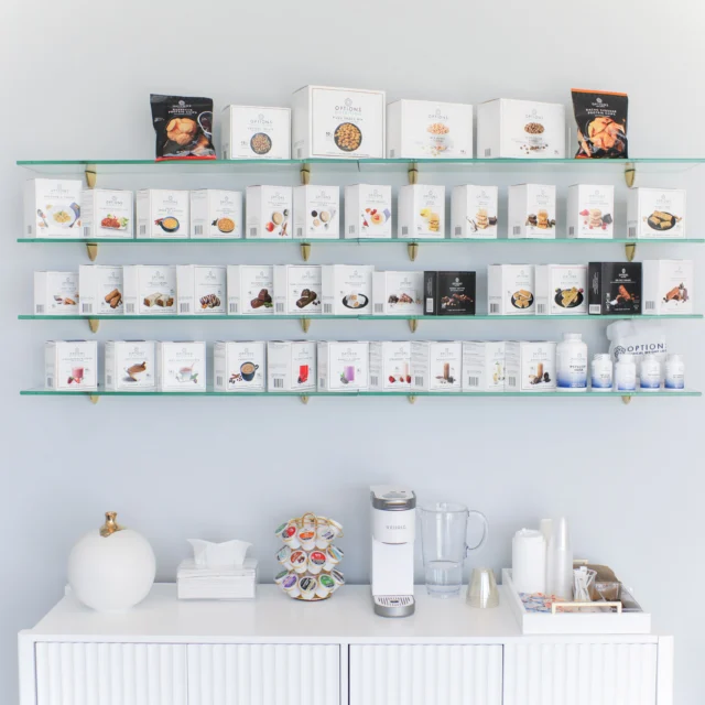 A minimalist coffee station with a coffee maker, creamers, cups, and tissues. Above are shelves displaying various packaged coffee products.