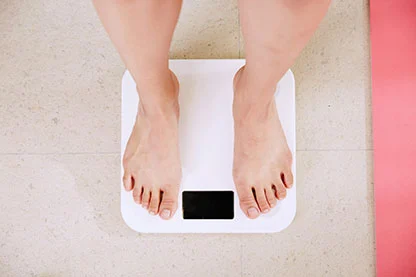 Person standing on a white digital bathroom scale, showing their bare feet. The scale's display is turned off or not visible. Flooring is tiled.