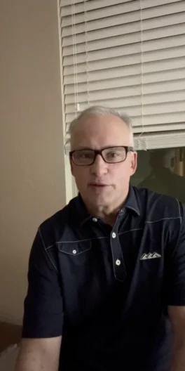 A man with short gray hair and glasses, wearing a dark polo shirt, sits indoors in front of a window with closed blinds.
