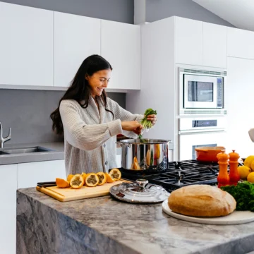 A woman in a kitchen adds greens to a pot on the stove. The counter has a cutting board with sliced squash, lemons, bread, and a red pot. White cabinets and an oven are in the background.