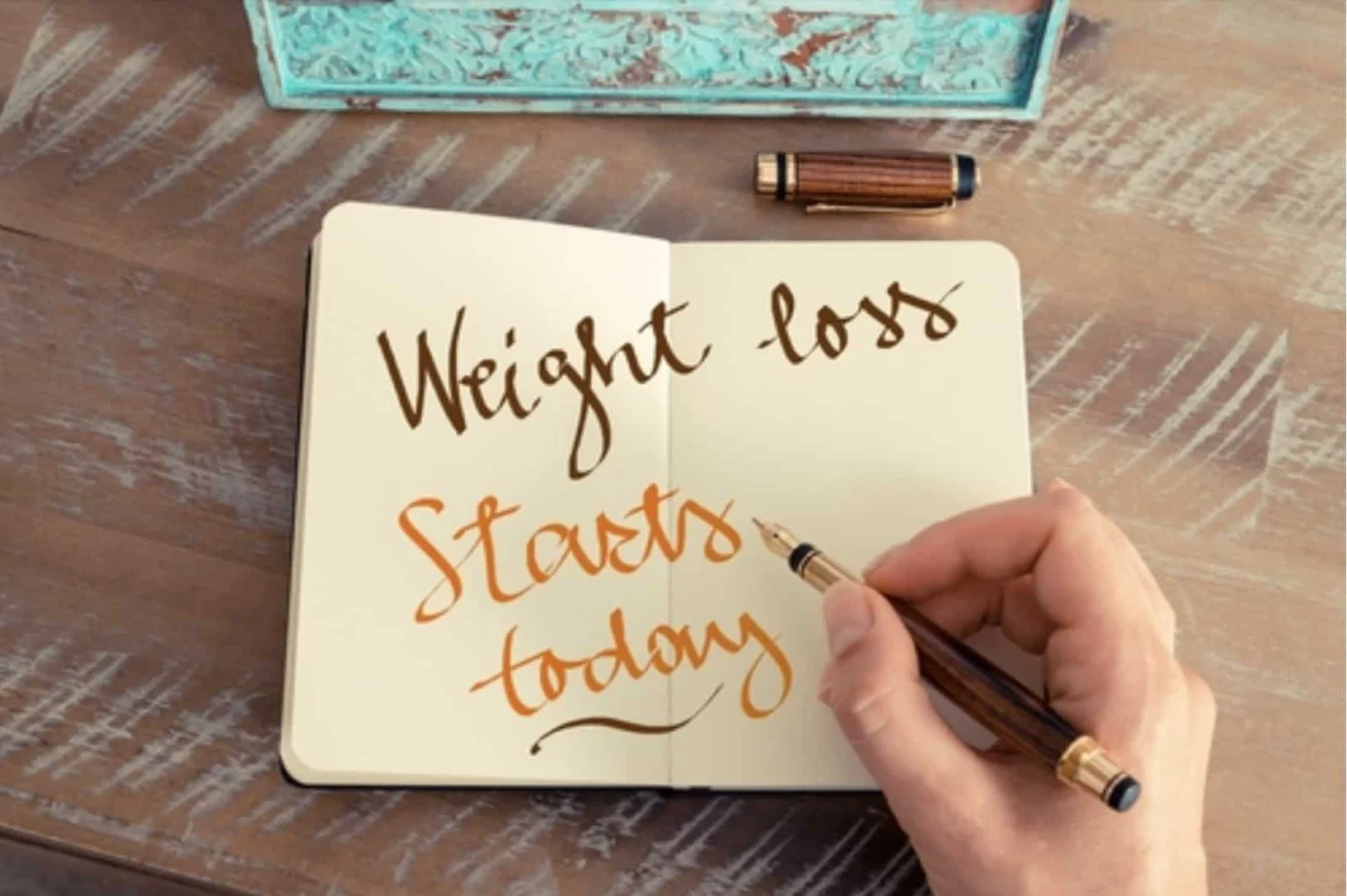 A hand writes "Weight loss starts today" in a notebook with a fountain pen on a wooden table.