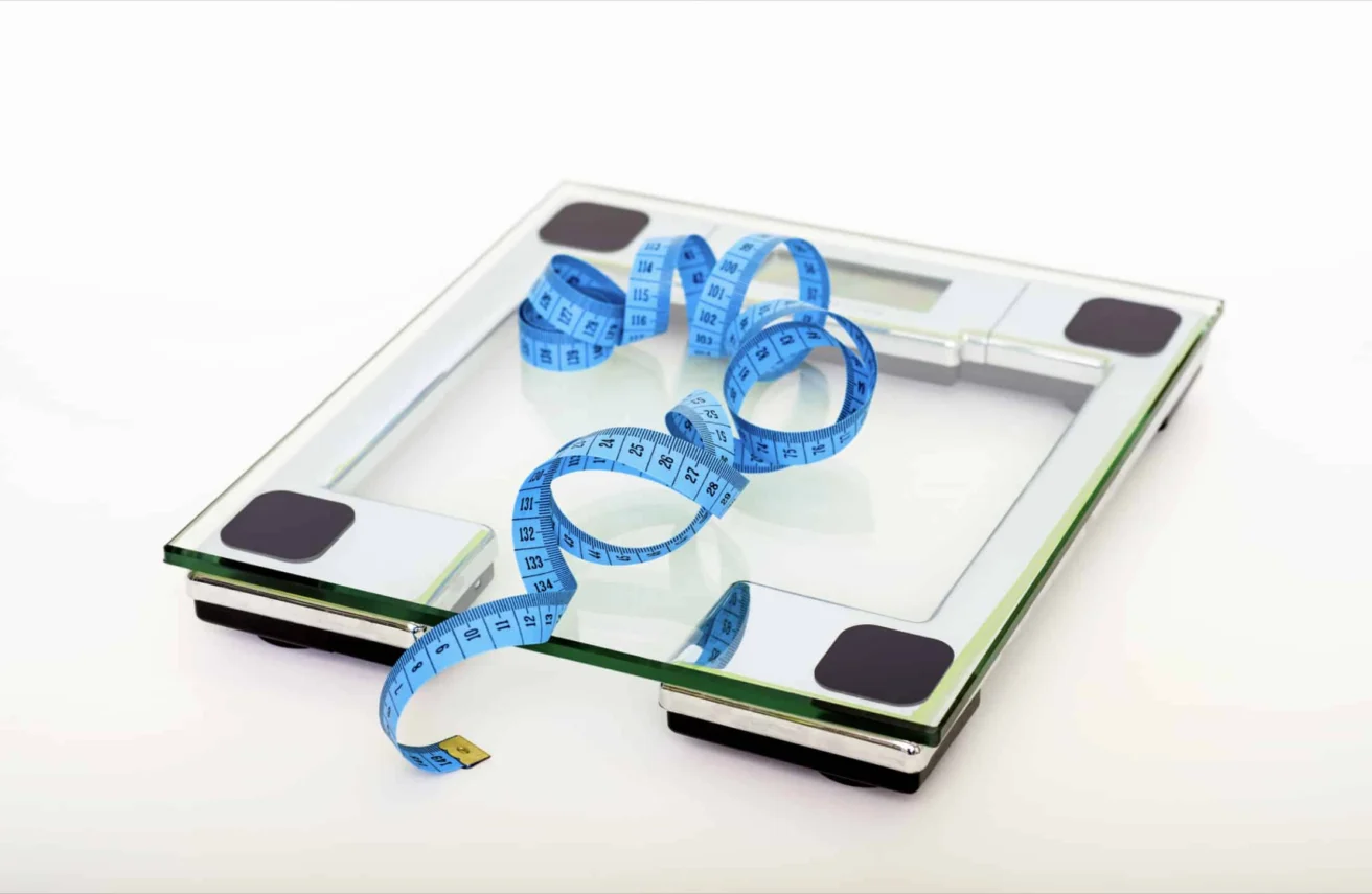 A glass digital bathroom scale with a blue measuring tape placed loosely on top, set against a white background.