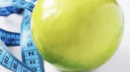 A green apple is placed on a white surface with a blue measuring tape coiled around it.