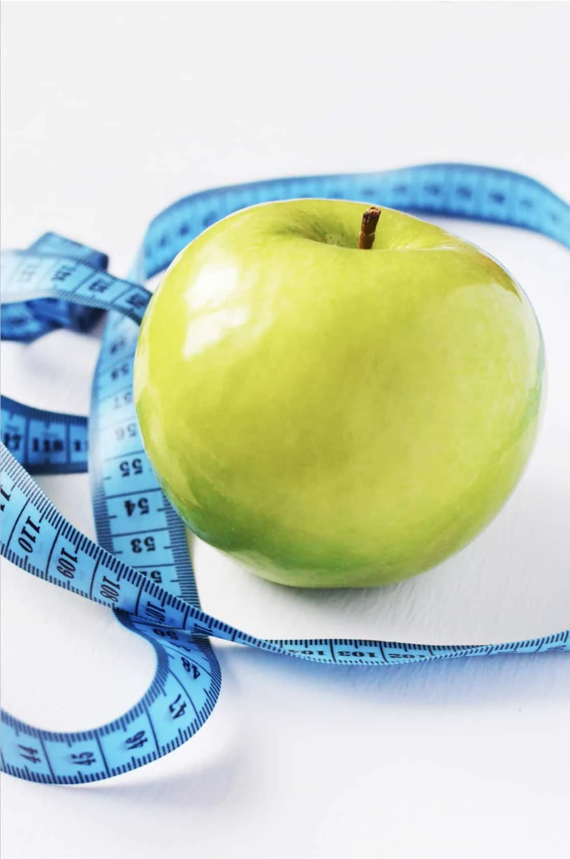 A green apple is placed on a white surface with a blue measuring tape coiled around it.