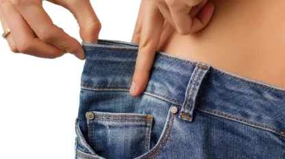 A person wearing loose blue jeans pulls the waistband away from their body, showing significant extra space around the waist.