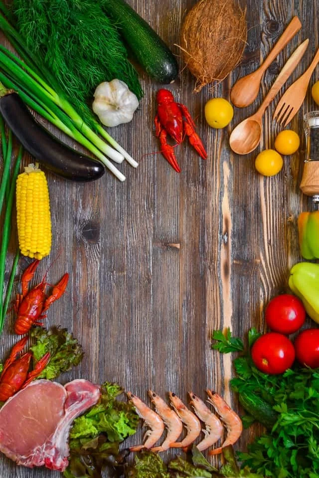 A variety of fresh vegetables, herbs, seafood, and meat are arranged on a rustic wooden table, forming a border around the edges and leaving the center empty.