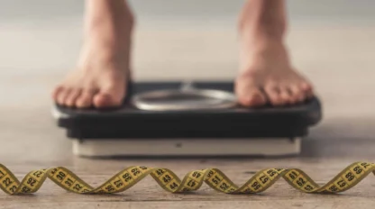 Person standing on a bathroom scale with a yellow measuring tape placed in the foreground.
