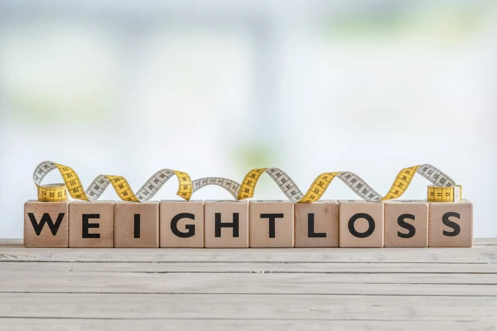 Wooden blocks spell "WEIGHT LOSS" on a wooden surface, with a yellow and white measuring tape coiled behind them.