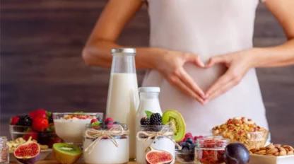 A person stands behind a table with hands forming a heart over their stomach. On the table are jars of yogurt, milk, fresh fruits, granola, nuts, and bowls of colorful berries, representing healthy eating.