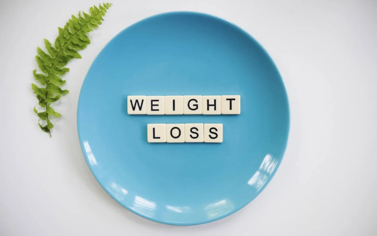 A blue plate with the words "WEIGHT LOSS" spelled out in letter tiles, next to a green fern leaf on a white background.
