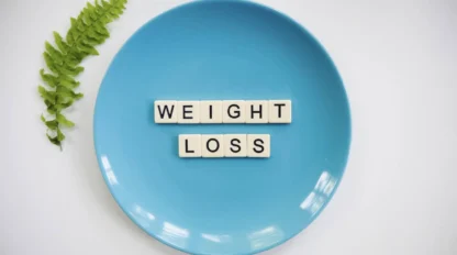 A blue plate with the words "WEIGHT LOSS" spelled out in letter tiles, next to a green fern leaf on a white background.