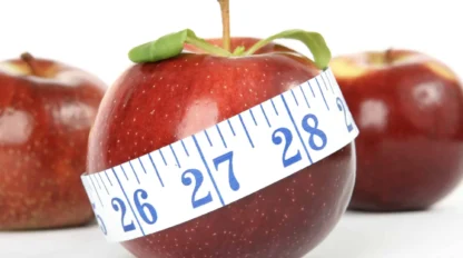 A red apple wrapped with a blue measuring tape, with two more apples in the background, against a white surface.