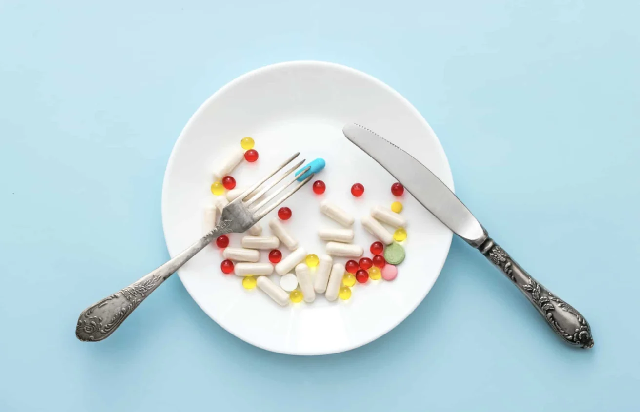 A white plate on a blue surface holds various pills and capsules, with a fork and knife placed on either side as if ready to eat the pills.