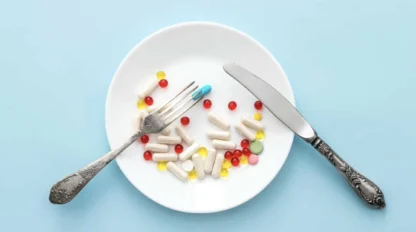 A white plate on a blue surface holds various pills and capsules, with a fork and knife placed on either side as if ready to eat the pills.