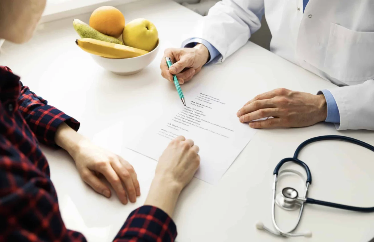 A doctor and patient review a medical form at a desk with a stethoscope and a bowl of fruit.
