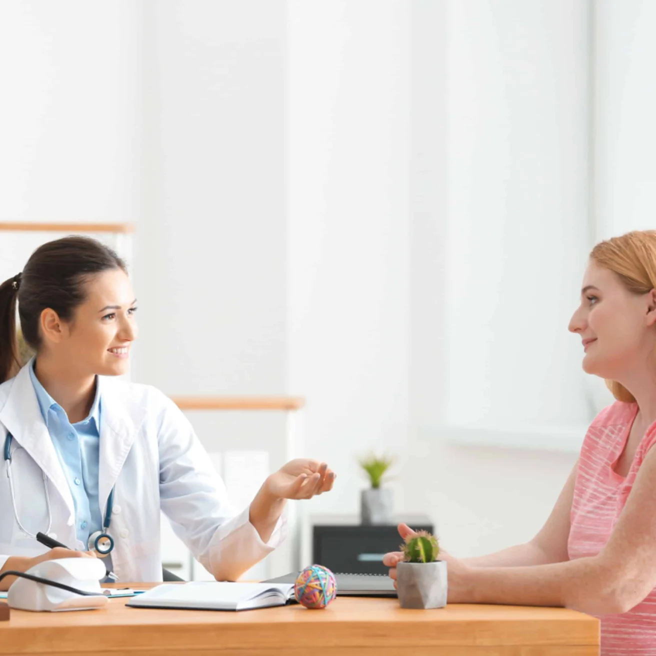 A doctor in a white coat sits at a desk and talks with a patient in a pink shirt across from her in a bright, modern office.