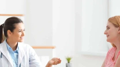 A doctor in a white coat sits at a desk and talks with a patient in a pink shirt across from her in a bright, modern office.