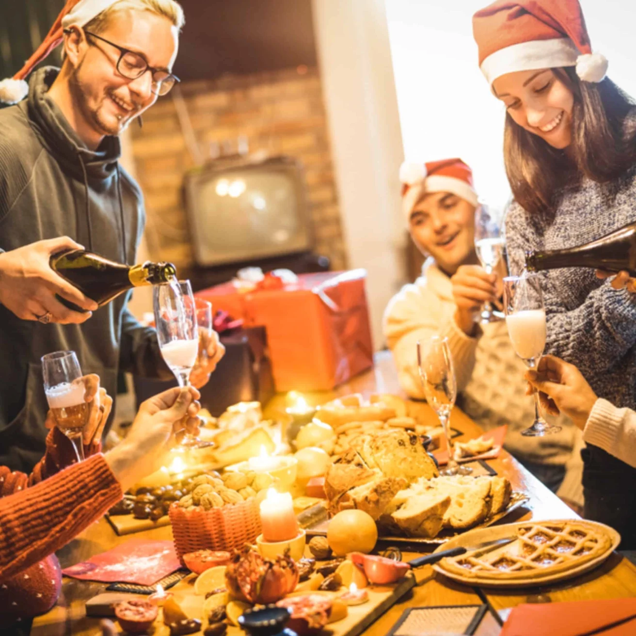 Four people in festive hats gather around a table with food, pouring drinks and celebrating, with gifts and candles in the background.