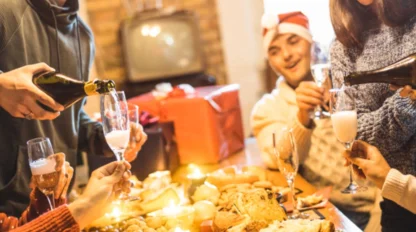 Four people in festive hats gather around a table with food, pouring drinks and celebrating, with gifts and candles in the background.
