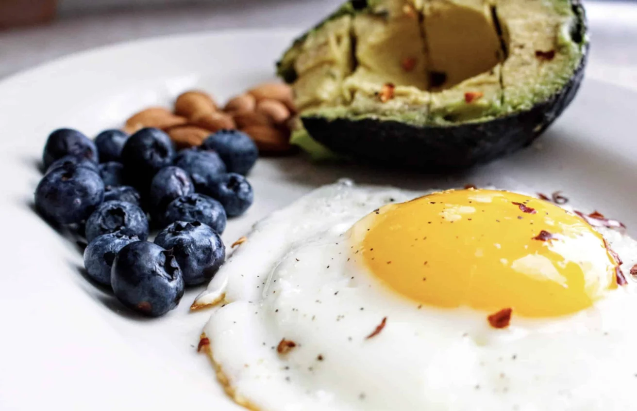 A plate with a sunny-side-up egg, half an avocado, a handful of almonds, and a small pile of blueberries.