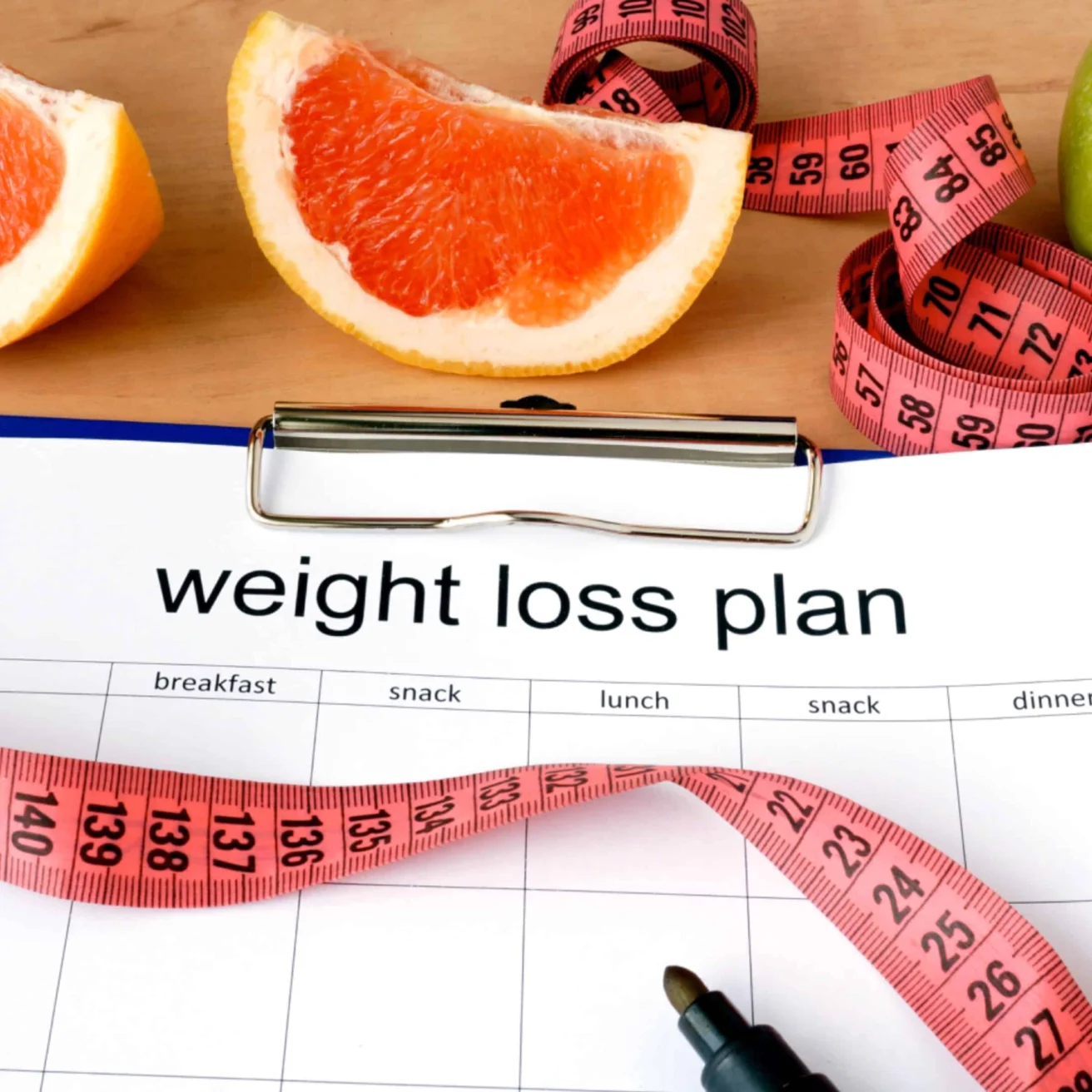 A clipboard with a weight loss plan chart, a pen, a pink measuring tape, and grapefruit slices on a wooden surface.