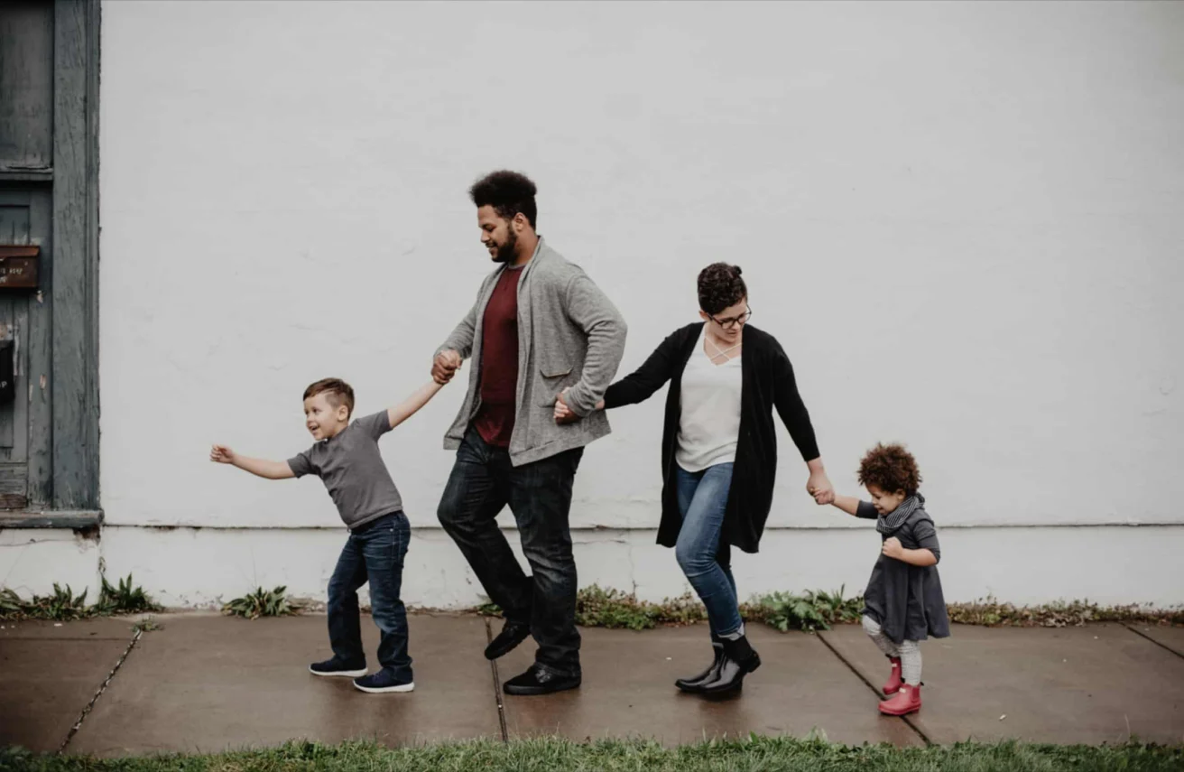 Four people, two adults and two children, hold hands and walk in a line on a sidewalk in front of a plain white wall.
