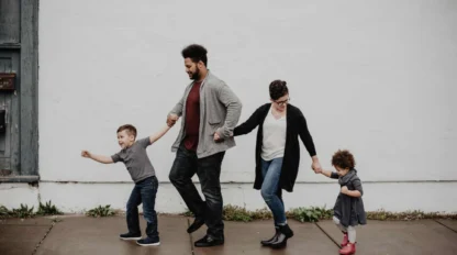 Four people, two adults and two children, hold hands and walk in a line on a sidewalk in front of a plain white wall.