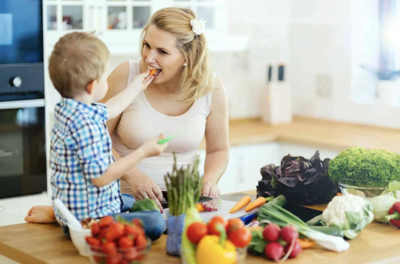 A young boy sits on a kitchen counter feeding a woman a carrot, surrounded by various fresh vegetables and fruits.