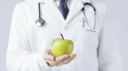 A person wearing a white lab coat and stethoscope holds out a green apple.