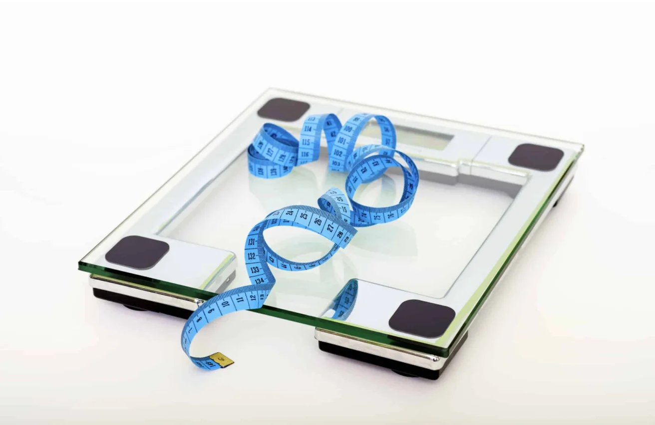 A digital glass bathroom scale with a blue measuring tape coiled on top, set against a plain white background.