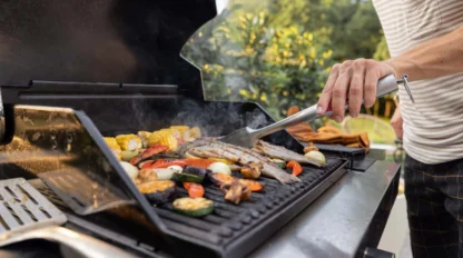 A person grills assorted vegetables and fish on an outdoor barbecue. Corn, bell peppers, zucchini, and onions are visible on the grill, with trees and greenery in the background.