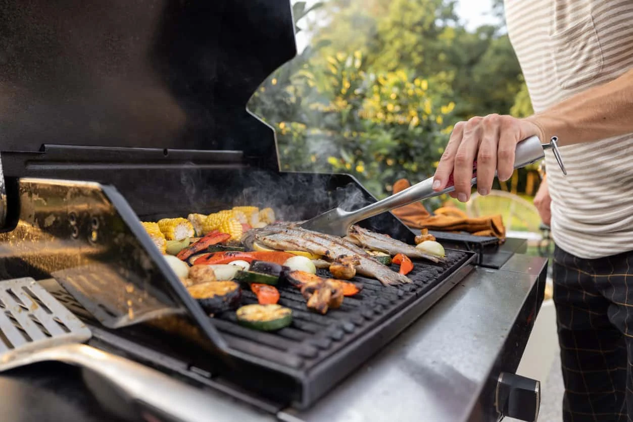 A person grills assorted vegetables and fish on an outdoor barbecue. Corn, bell peppers, zucchini, and onions are visible on the grill, with trees and greenery in the background.