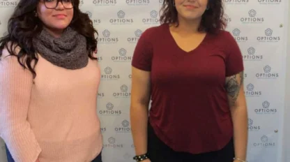 Two women stand side by side in front of a backdrop with "OPTIONS" logos. One wears a pink sweater and scarf; the other wears a red t-shirt. The date "04/25/2019" appears in the corner.