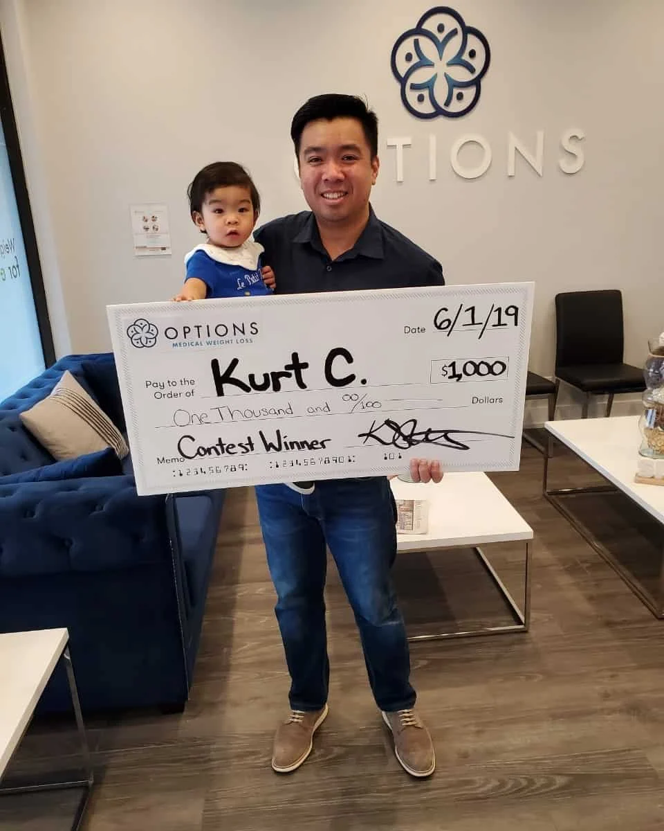 A man holds a toddler and a large check for $1,000 awarded to "Kurt C." as a contest winner, dated 6/1/19, inside an office with "OPTIONS" on the wall.