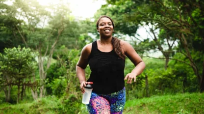 A woman in colorful leggings and a black tank top jogs outdoors on a grassy path, holding a water bottle and smiling, surrounded by lush green trees and plants.