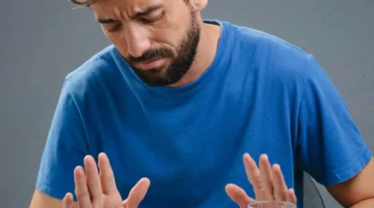A man in a blue shirt looks uncomfortable and pushes away a plate of food with tomato sauce, with a glass of water on the table.