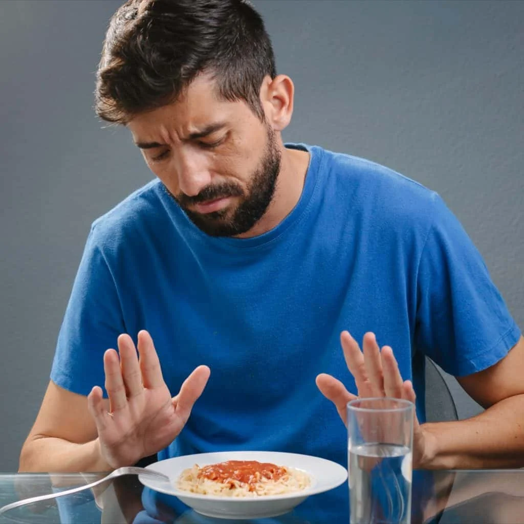 A man in a blue shirt looks uncomfortable and pushes away a plate of food with tomato sauce, with a glass of water on the table.