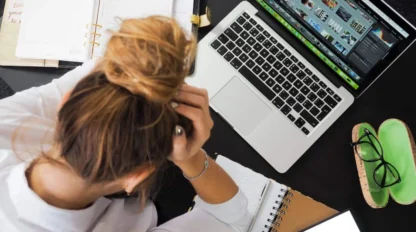 A person with their head in their hands sits at a desk with a laptop, open notebooks, a smartphone, and glasses case.
