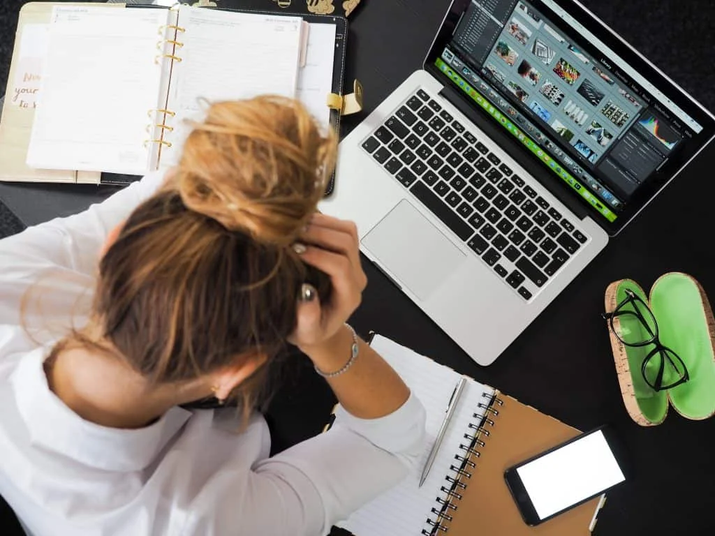 A person with their head in their hands sits at a desk with a laptop, open notebooks, a smartphone, and glasses case.