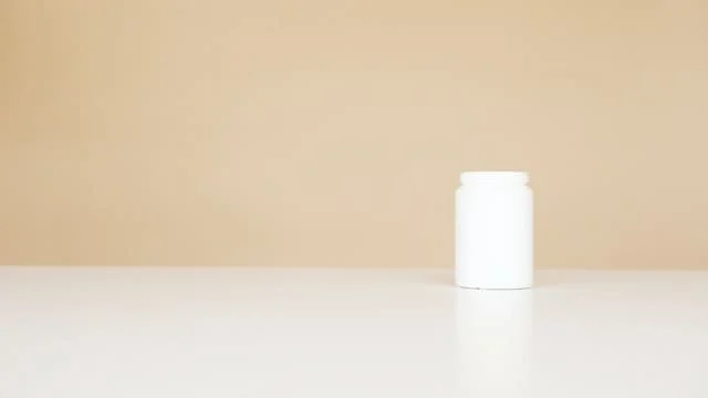 A small, plain white plastic jar with a lid sits on a white surface against a beige background. The container is centered to the right side of the image, and the scene is minimalistic.